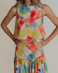 A woman wearing a round neck sleeveless blouse that is mostly yellow with other watercolors.