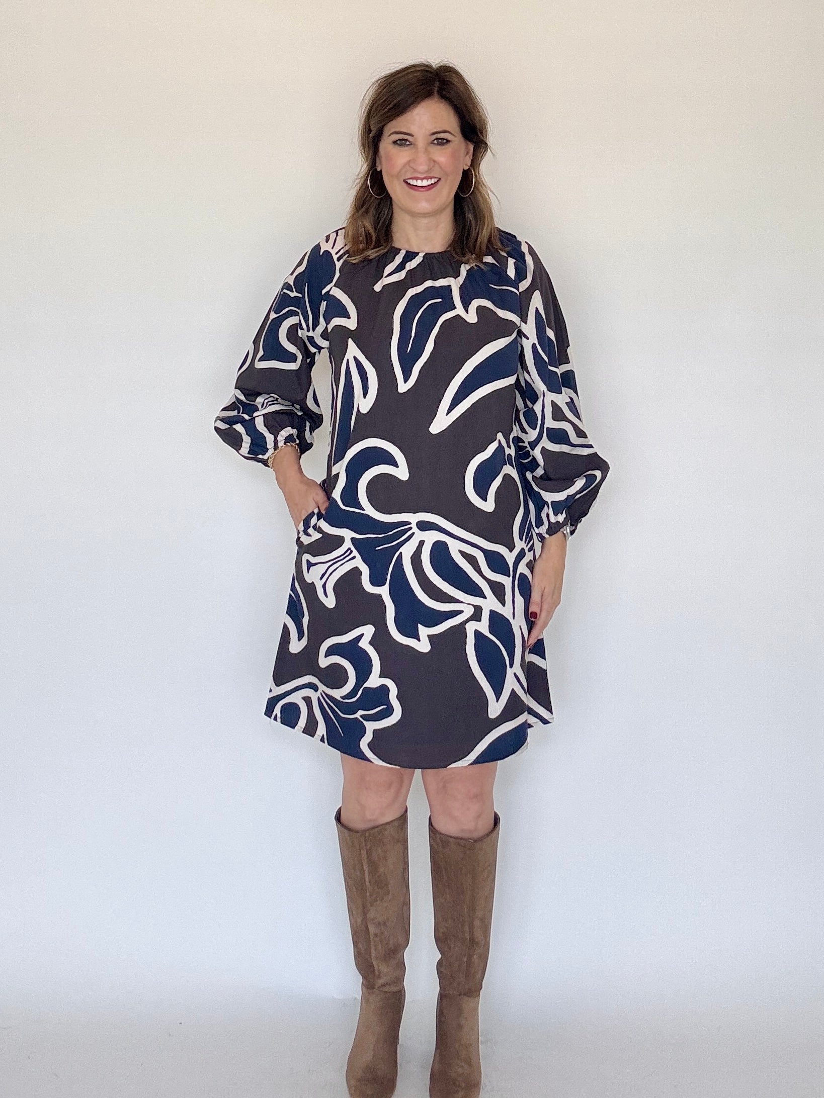 A woman modeling a navy blue and white floral print shift dress with three-quarter sleeves and pockets, paired with brown knee-high boots, standing against a plain white wall.