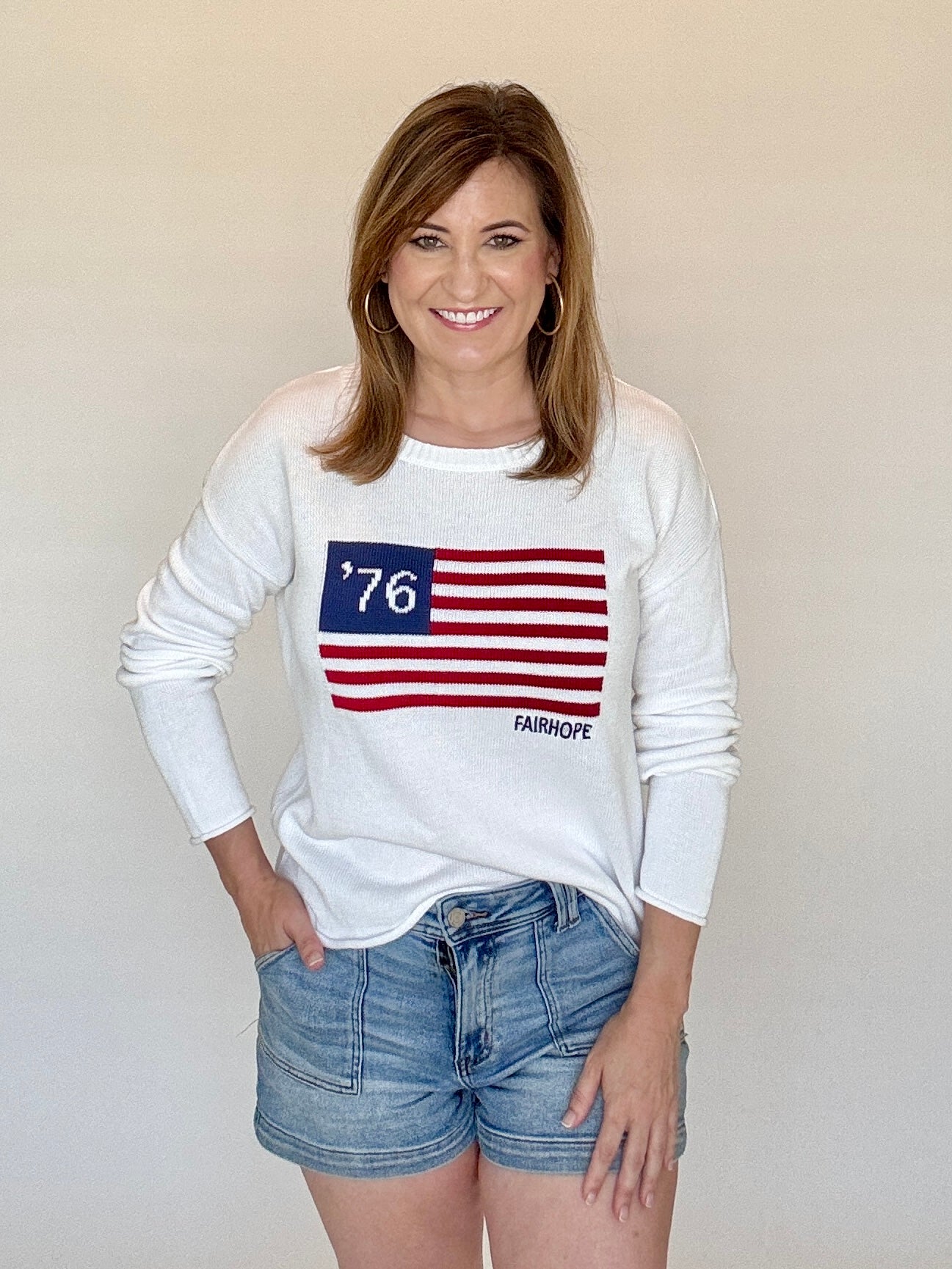 Woman wearing a white sweatshirt with an American flag design and denim shorts against a plain background