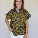 A woman wearing a short-sleeve button-up blouse with an olive green and black leopard print, paired with white pants, standing against a plain light beige background.