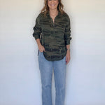 A woman stands against a plain off-white wall wearing a long-sleeve camouflage button-up shirt, light blue jeans, and brown woven shoes.