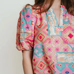 A woman wearing a brightly colored puff sleeve v-neck blouse with oranges, pinks and greens.