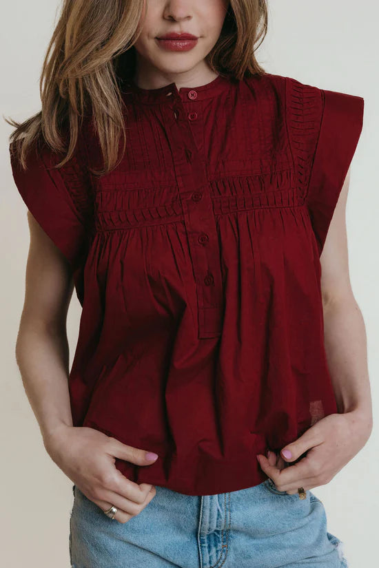 A woman wearing a garnet colored sleeveless blouse with pleats along the collar.