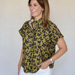 A woman modeling a short-sleeve button-up blouse with an olive green and black floral pattern, paired with white pants, against a plain light beige background.