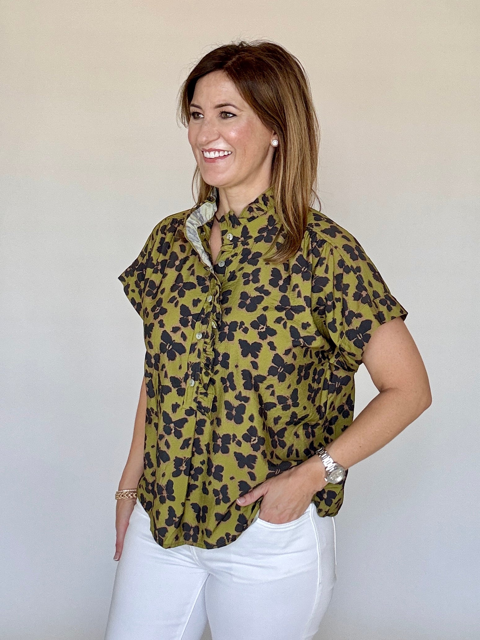 A woman modeling a short-sleeve button-up blouse with an olive green and black floral pattern, paired with white pants, against a plain light beige background.