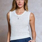 Woman wearing a white sleeveless crochet top and blue pants against a neutral background