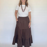 A woman modeling a brown tiered maxi skirt with a white short-sleeve top and layered necklaces, standing against a plain off-white background.