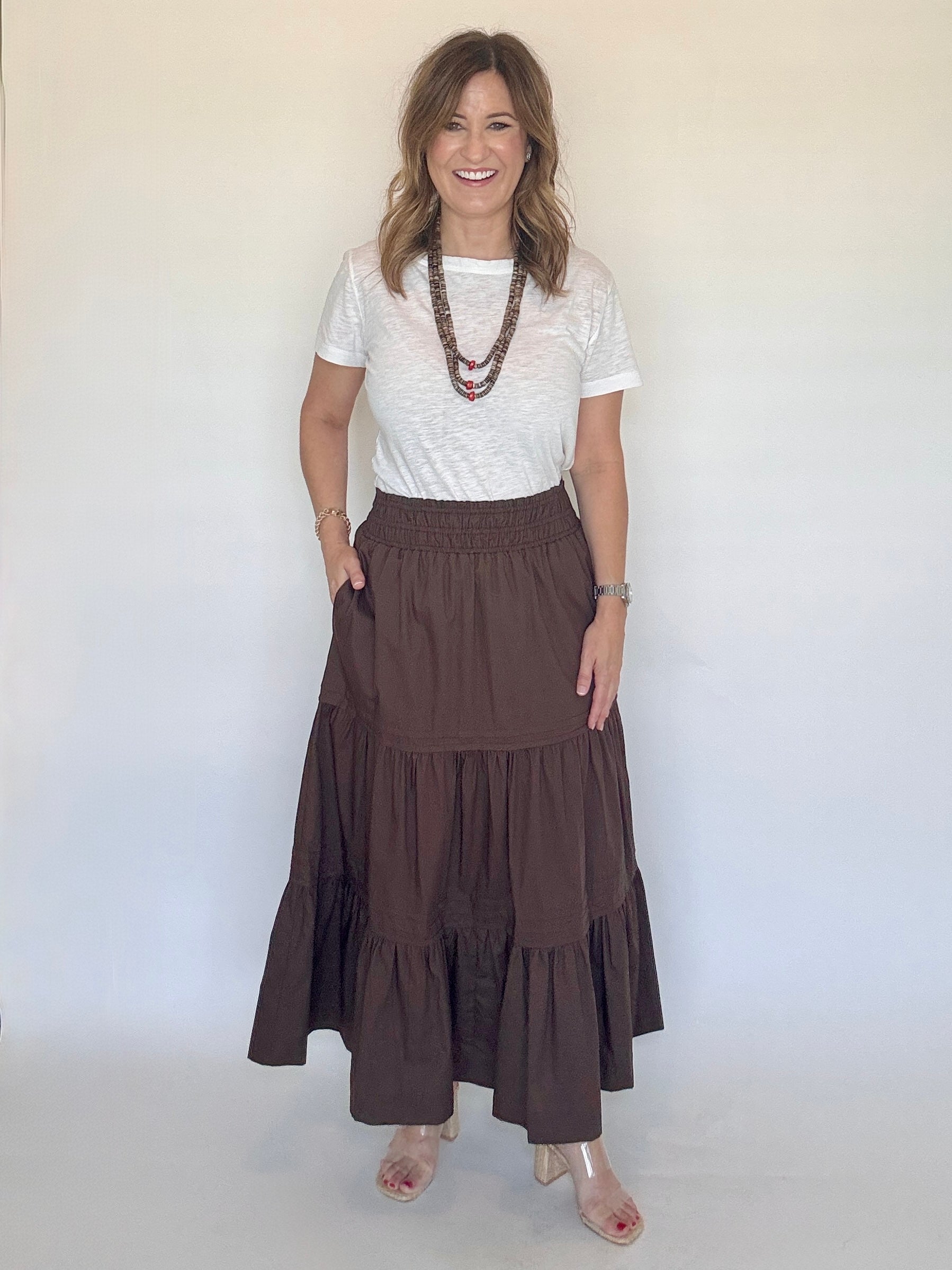 A woman modeling a brown tiered maxi skirt with a white short-sleeve top and layered necklaces, standing against a plain off-white background.