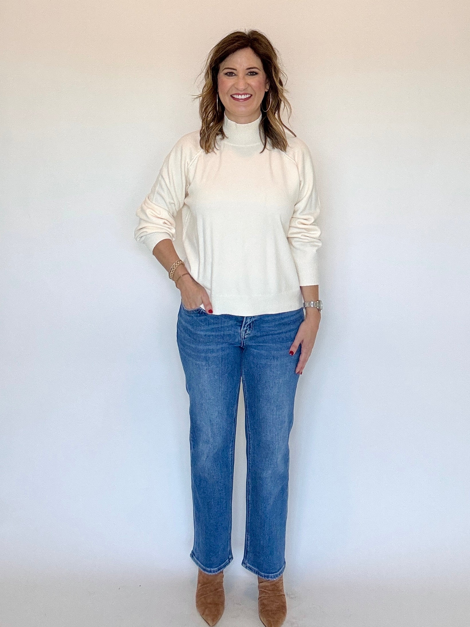 A woman modeling a cream-colored long-sleeve mock neck sweater paired with blue jeans and brown suede boots, standing against a plain white studio background.