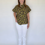 A woman modeling a short-sleeve button-up shirt with an olive green and black leopard print, paired with white pants and tan sandals, standing against a plain white background.