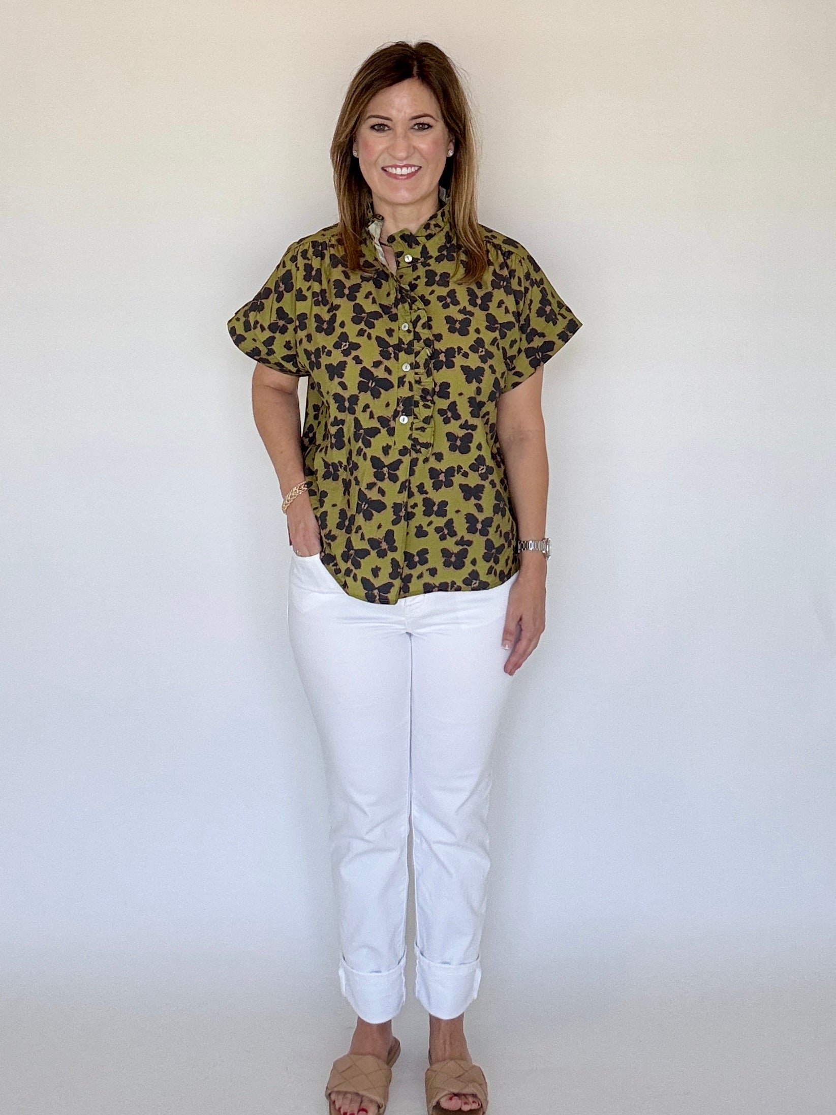 A woman modeling a short-sleeve button-up shirt with an olive green and black leopard print, paired with white pants and tan sandals, standing against a plain white background.