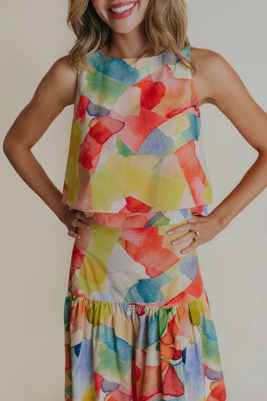 A woman wearing a round neck sleeveless blouse that is mostly yellow with other watercolors.
