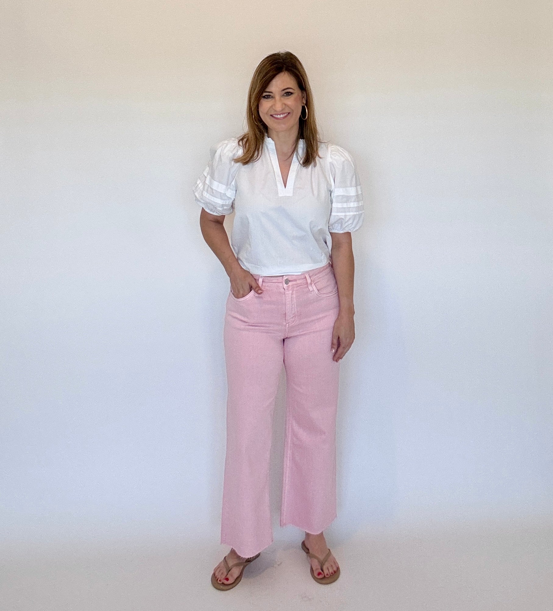 Woman wearing a white blouse and pink wide leg ankle crop jeans  against a plain background