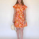 A woman wearing an orange, pink and yellow floral mini shirtdress with a collar and a belt.
