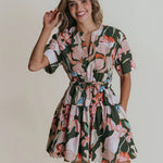 A woman wearing a split neck mini dress with a cinched tie waist in a green floral print.