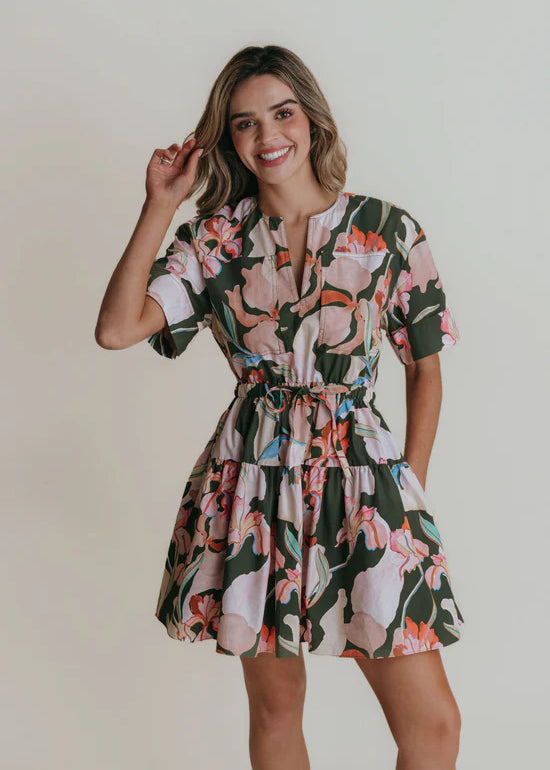A woman wearing a split neck mini dress with a cinched tie waist in a green floral print.
