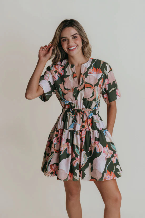A woman wearing a split neck mini dress with a cinched tie waist in a green floral print.