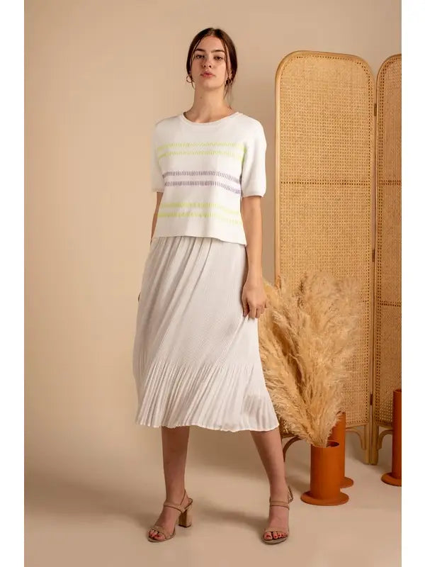 Woman wearing a white top and pleated skirt standing next to a woven screen indoors.
