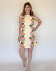 A woman wearing a cream print mini dress that has pink and yellow floral borders along the side and buttons up and down the sides of the dress.