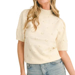 A woman wearing a cream-colored short-sleeve knit top with small raised pearl-like embellishments, paired with light-wash blue jeans, against a plain beige studio background.