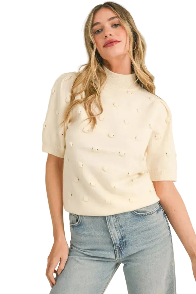 A woman wearing a cream-colored short-sleeve knit top with small raised pearl-like embellishments, paired with light-wash blue jeans, against a plain beige studio background.