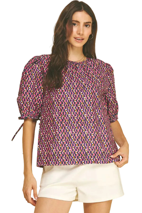 A model modeling the navy and pink geometric print puff sleeve blouse by PINCH with self-tie cuff detail, paired with white shorts, from Chatsworth Boutique.