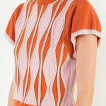A model posing in the Orange Ruffle Knit Blouse by THML, a soft knit blouse in a wavy orange, pink, and white print, from Chatsworth Boutique in Fairhope, Alabama.
