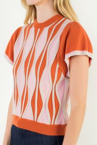 A model posing in the Orange Ruffle Knit Blouse by THML, a soft knit blouse in a wavy orange, pink, and white print, from Chatsworth Boutique in Fairhope, Alabama.